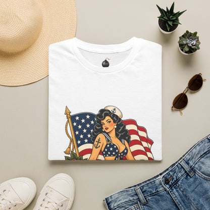 A folded white graphic T-shirt is displayed in a styled flat lay on a light beige surface, surrounded by a straw hat, small potted succulents, sunglasses, white sneakers, and blue jeans. The shirt features a retro pin-up woman graphic with an American flag and gold anchor details.