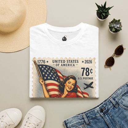 A folded white graphic T-shirt is styled in a flat lay on a light beige surface, surrounded by a straw hat, white sneakers, blue jeans, sunglasses, and small potted succulents. The shirt features a vintage-style postage stamp design with an American flag, a smiling woman, and the text '1776 United States of America 2026 78c U.