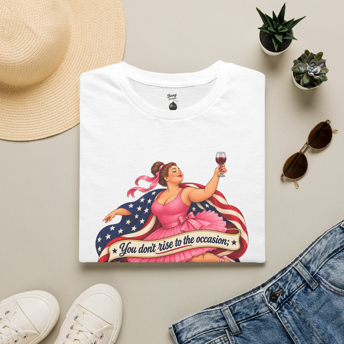A folded white graphic T-shirt is displayed in a styled flat lay on a light beige surface, surrounded by a straw hat, white sneakers, blue jeans, small potted succulents, and sunglasses. The shirt features a retro-style illustration of a woman in a pink dress holding a wine glass, with an American flag motif and the text 'You don't rise to the occasion.